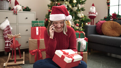 Obraz na płótnie Canvas Young blonde woman talking on smartphone sitting on floor by christmas tree at home