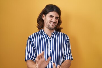 Hispanic man with long hair standing over yellow background disgusted expression, displeased and...