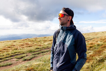 young man in jacket and sunglasses stands on the mountain and looks at the sun, tourist guy in...