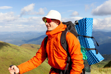young man in orange jacket and glasses stands on the top of mountain with backpack and trekking...