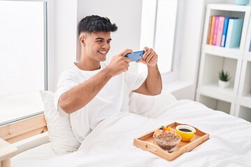 Young hispanic man make photo by smartphone to breakfast at bedroom