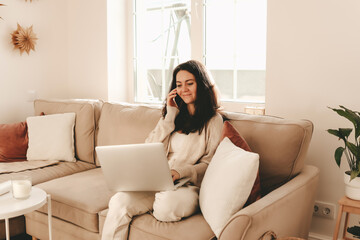 A young businesswoman in casual clothes works online from home using a laptop and wireless technology while sitting on a sofa in a cozy room at home. Selective focus