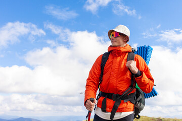 young man in orange jacket and glasses walks in the mountains with trekking stick, tourist guy with...