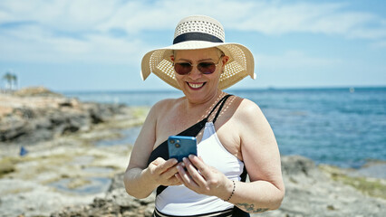 Fototapeta na wymiar Middle age grey-haired woman tourist wearing swimsuit using smartphone at the beach
