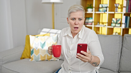 Middle age grey-haired woman using smartphone drinking coffee with surprise expression at home