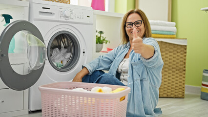 Middle age hispanic woman smiling confident washing clothes doing thumb up gesture at laundry room