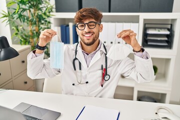 Arab man with beard wearing doctor uniform holding medical mask winking looking at the camera with...