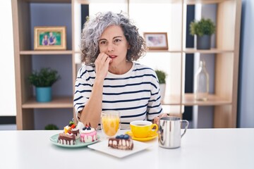 Middle age woman with grey hair eating pastries and drinking coffee for breakfast looking stressed and nervous with hands on mouth biting nails. anxiety problem.
