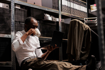 Private investigator drinking coffee and using laptop on floor in dark agency office. Police detective analyzing crime case on computer, searching crime case solving solution at night