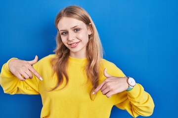 Young caucasian woman standing over blue background looking confident with smile on face, pointing...