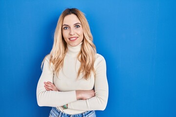 Young caucasian woman standing over blue background happy face smiling with crossed arms looking at the camera. positive person.