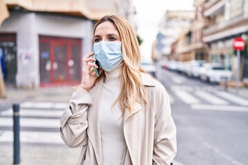 Young blonde woman wearing medical mask talking on smartphone at street