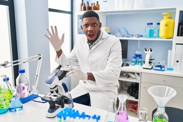 African american man working at scientist laboratory scared and amazed with open mouth for surprise, disbelief face