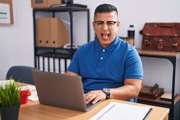 Young hispanic man working at the office with laptop sticking tongue out happy with funny expression. emotion concept.