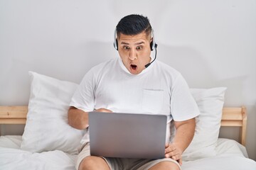 Hispanic young man using computer laptop on the bed scared and amazed with open mouth for surprise, disbelief face