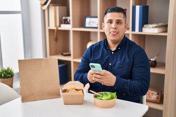 Hispanic young man eating take away food using smartphone sticking tongue out happy with funny expression.