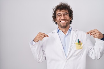 Hispanic young man wearing doctor uniform looking confident with smile on face, pointing oneself...