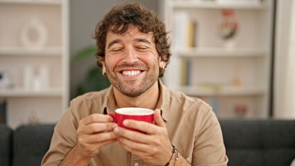 Young hispanic man smelling cup of coffee sitting on sofa smiling at home