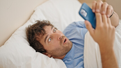 Fototapeta na wymiar Young hispanic man using smartphone lying on bed surprised at bedroom