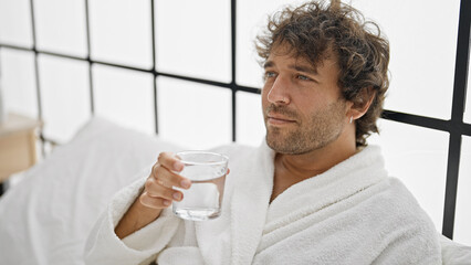 Young hispanic man wearing bathrobe drinking water at bedroom