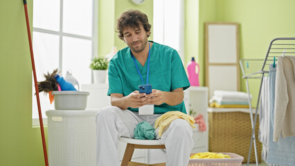 Young hispanic man professional cleaner using smartphone relaxing at laundry room
