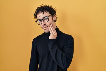 Hispanic man standing over yellow background touching mouth with hand with painful expression because of toothache or dental illness on teeth. dentist