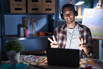 Young hispanic man sitting at art studio with laptop late at night smiling looking to the camera showing fingers doing victory sign. number two.