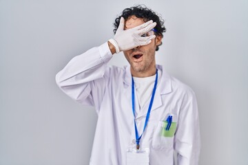 Hispanic man working at scientist laboratory peeking in shock covering face and eyes with hand, looking through fingers afraid