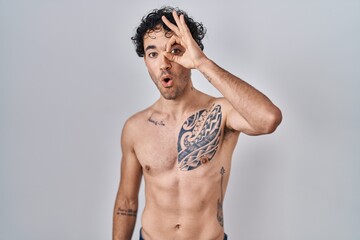 Fototapeta premium Hispanic man standing shirtless doing ok gesture shocked with surprised face, eye looking through fingers. unbelieving expression.