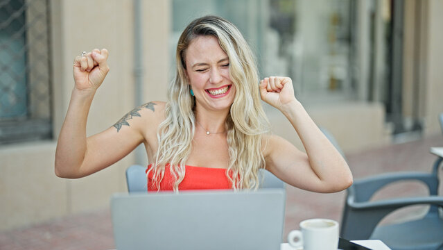 Young blonde woman using laptop sitting on table with winner gesture at coffee shop terrace