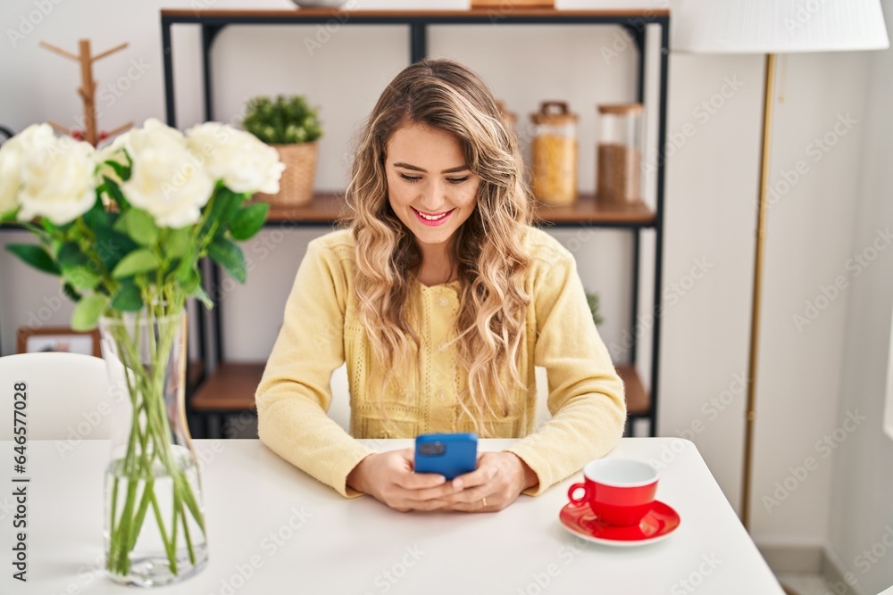 Wall mural Young woman drinking coffee using smartphone at home