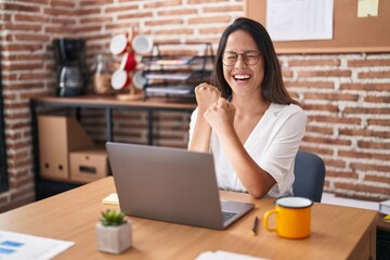 Hispanic young woman working at the office wearing glasses celebrating surprised and amazed for success with arms raised and eyes closed