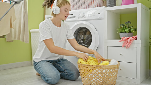 Young blonde woman listening to music holding clothes of basket at laundry room
