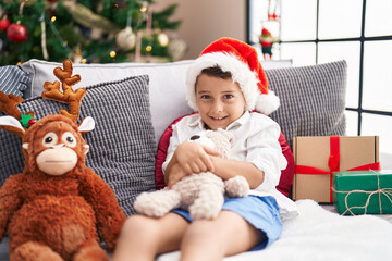 Adorable hispanic toddler hugging teddy bear sitting on sofa by christmas tree at home