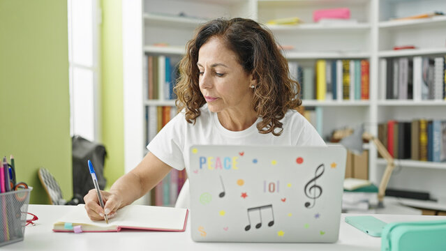 Middle Age Hispanic Woman Student Using Laptop Writing On Notebook At Library University