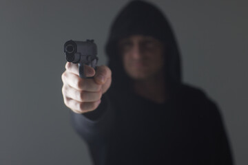 robber with gun aiming into camera. Man in hood threatens with firearm. Weapon in person's hands....