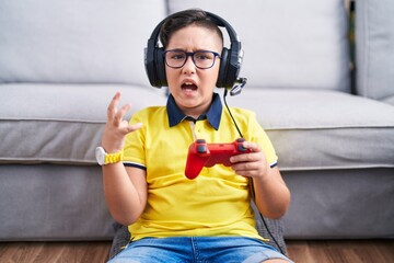 Young hispanic kid playing video game holding controller wearing headphones crazy and mad shouting...