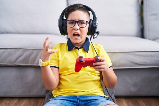 Young Hispanic Kid Playing Video Game Holding Controller Wearing Headphones Crazy And Mad Shouting And Yelling With Aggressive Expression And Arms Raised. Frustration Concept.