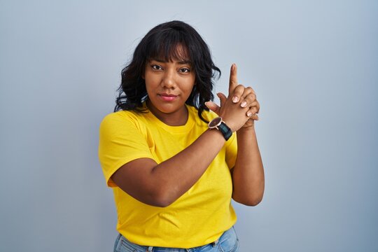 Hispanic woman standing over blue background holding symbolic gun with hand gesture, playing killing shooting weapons, angry face