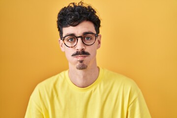 Hispanic man wearing glasses standing over yellow background puffing cheeks with funny face. mouth inflated with air, crazy expression.