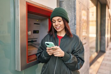 Young beautiful hispanic woman using smartphone standing by bank teller at street
