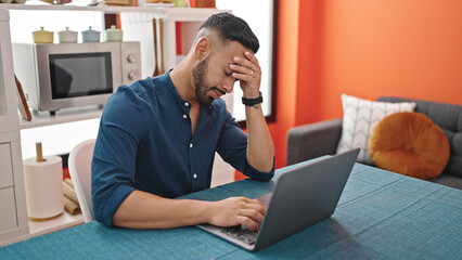 Young hispanic man stressed using laptop sitting on the table at dinning room