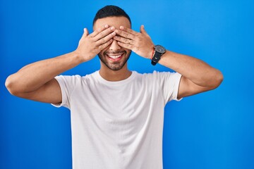 Young hispanic man standing over blue background covering eyes with hands smiling cheerful and...