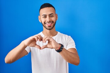 Young hispanic man standing over blue background smiling in love doing heart symbol shape with hands. romantic concept.