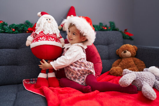 Adorable blonde toddler playing with santa claus doll sitting on sofa by christmas decoration at home