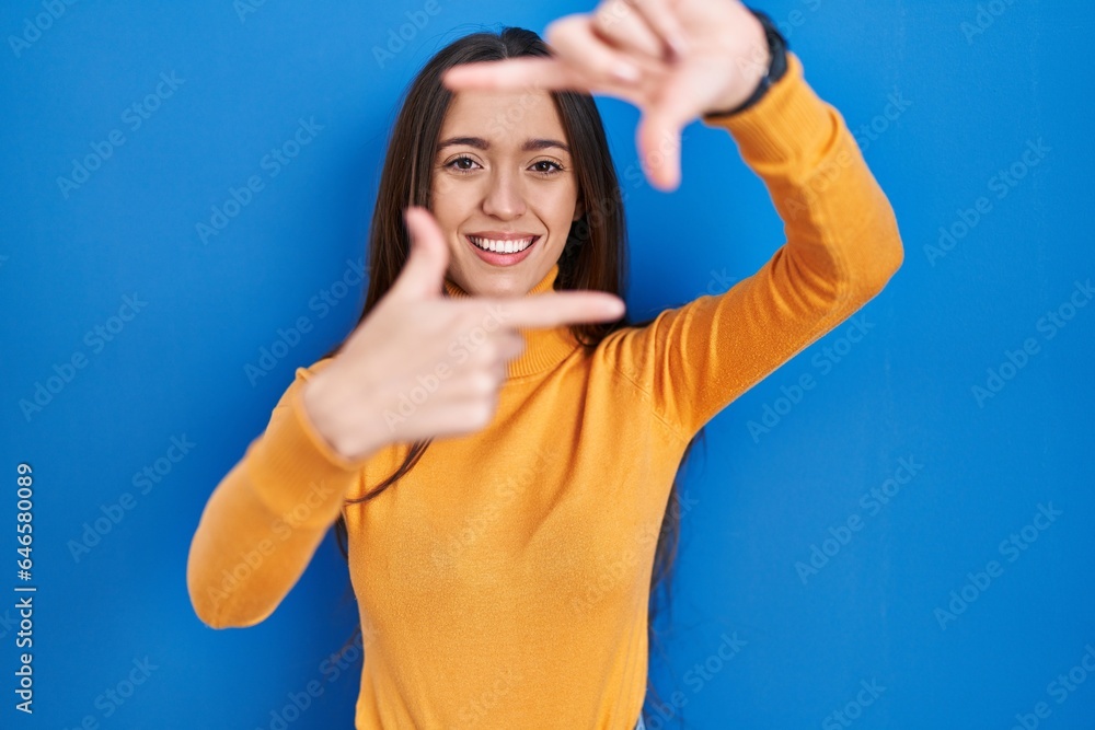 Sticker young brunette woman standing over blue background smiling making frame with hands and fingers with 