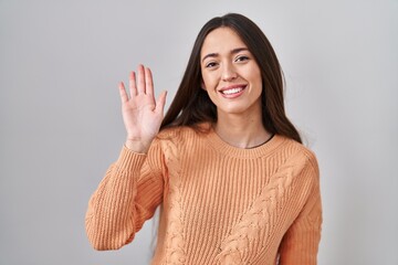 Young brunette woman standing over white background waiving saying hello happy and smiling, friendly welcome gesture © Krakenimages.com