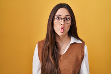 Young brunette woman standing over yellow background wearing glasses making fish face with lips,...