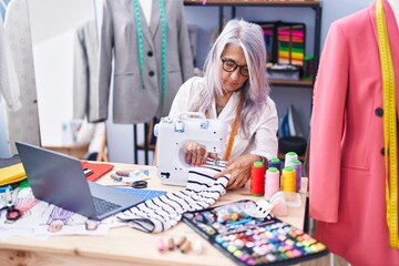 Fototapeta premium Middle age grey-haired woman tailor using sewing machine and laptop at tailor shop