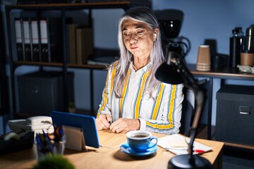 Middle age woman with grey hair working at the office at night puffing cheeks with funny face. mouth inflated with air, crazy expression. - obrazy, fototapety, plakaty